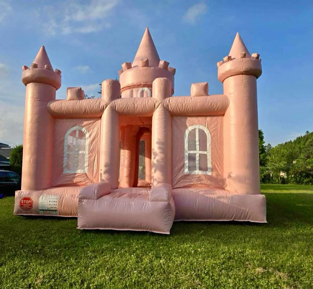 Château gonflable rose pâle à trois tours pointues, vu de face sur le gazon, sous un ciel bleu